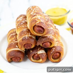 pretzel wrapped hot dogs stacked in a pyramid shape on white plate with bowl of mustard in the background.