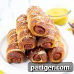 pretzel wrapped hot dogs stacked in a pyramid shape on white plate with bowl of mustard in the background.