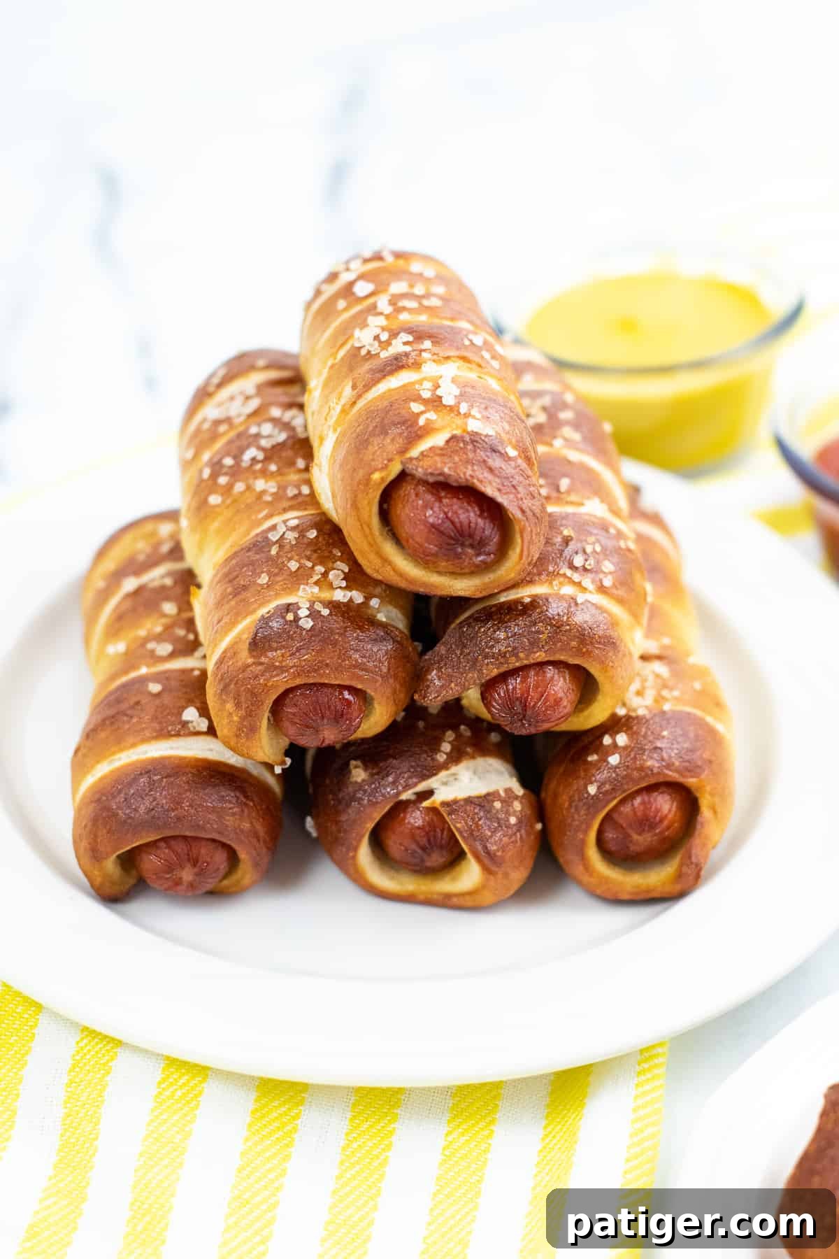 pretzel wrapped hot dogs stacked in a pyramid shape on white plate with bowl of mustard in the background.