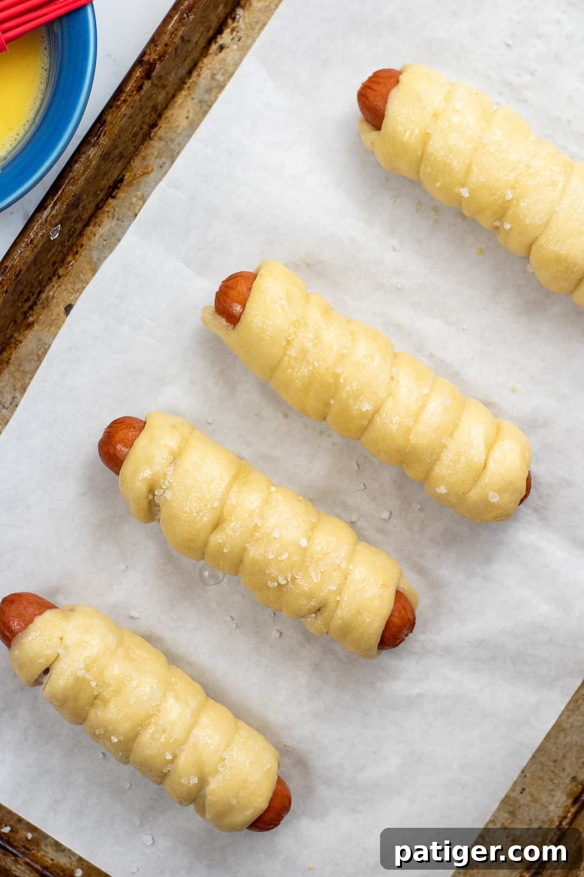 unbaked pretzel dough wrapped hot dogs with coarse salt on top on baking sheet.
