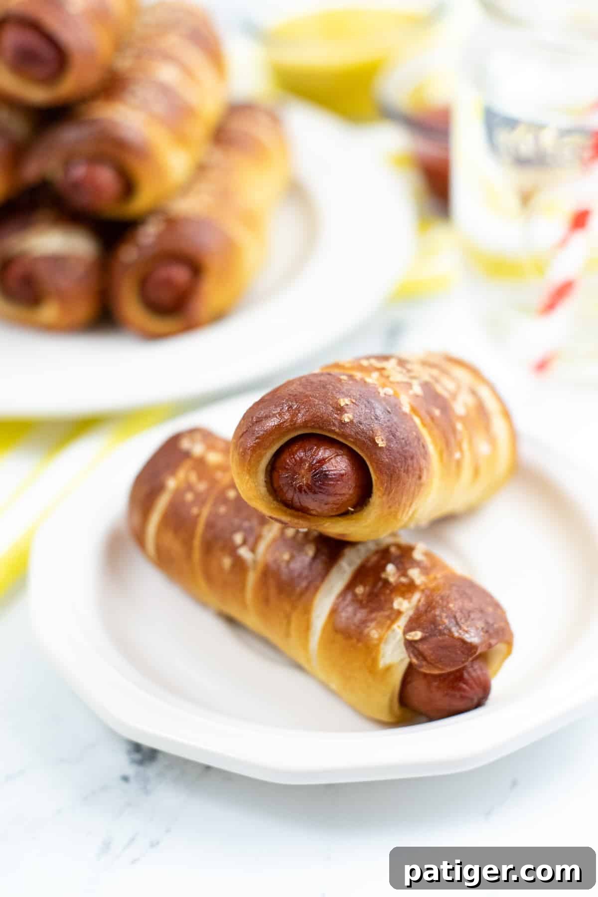 Two Pretzel Hot Dogs on white plate with more pretzel hot dogs on plate in background.