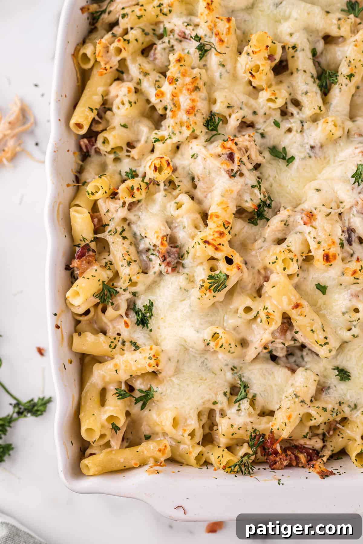 Golden Alfredo Chicken Bake 11 Corner of a baking dish filled with a cheesy chicken bacon alfredo pasta bake, garnished with fresh parsley, highlighting the rich, bubbly cheese.