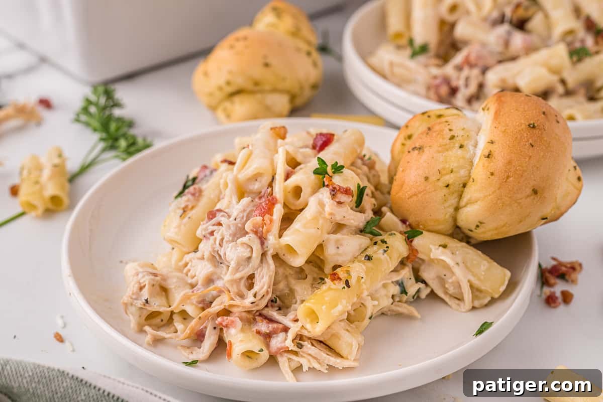 Golden Alfredo Chicken Bake 12 A serving of cheesy chicken alfredo pasta bake on a white plate, with a roll and additional plates blurred in the background, ready to be enjoyed.