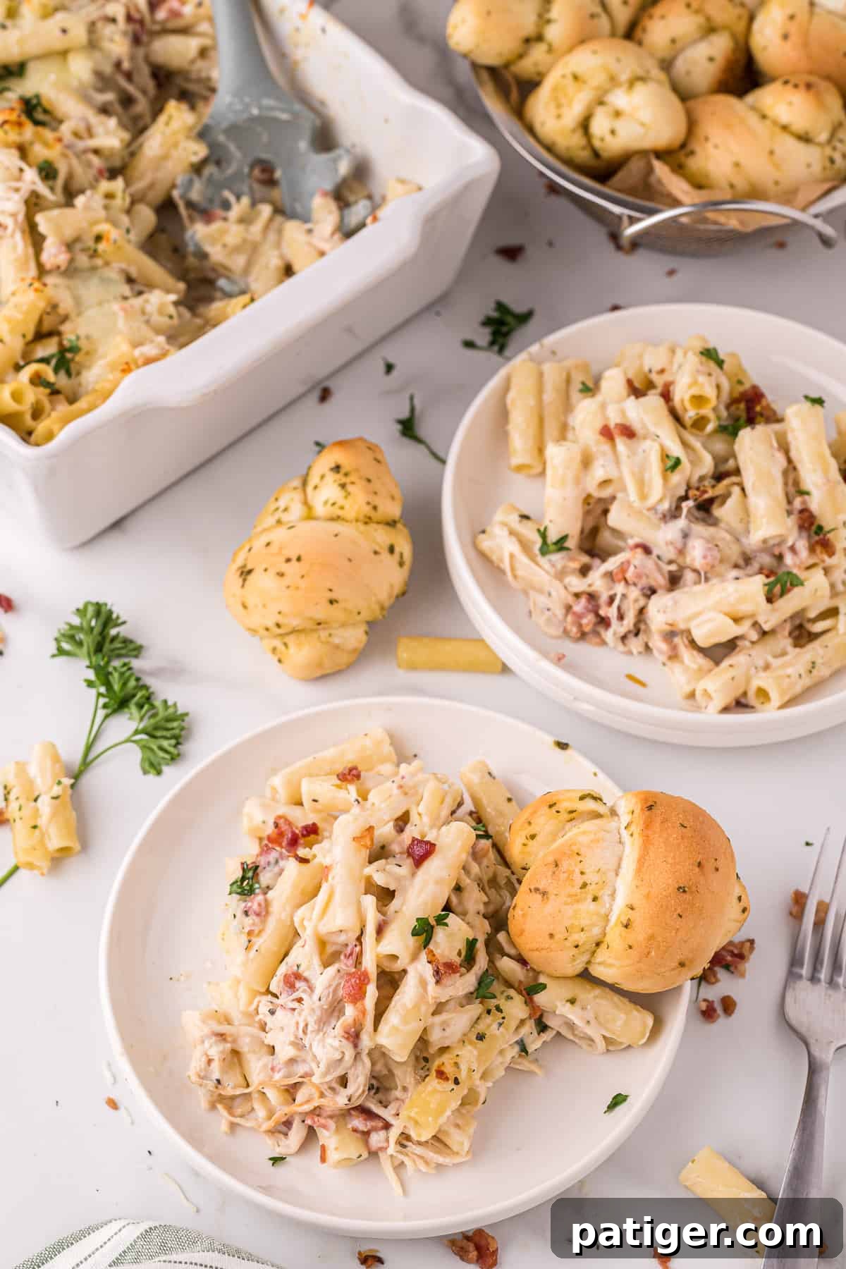 Golden Alfredo Chicken Bake 4 A serving of Chicken Bacon Ziti Pasta Bake on a white plate, accompanied by a garlic knot. Additional servings and the casserole dish are blurred in the background.