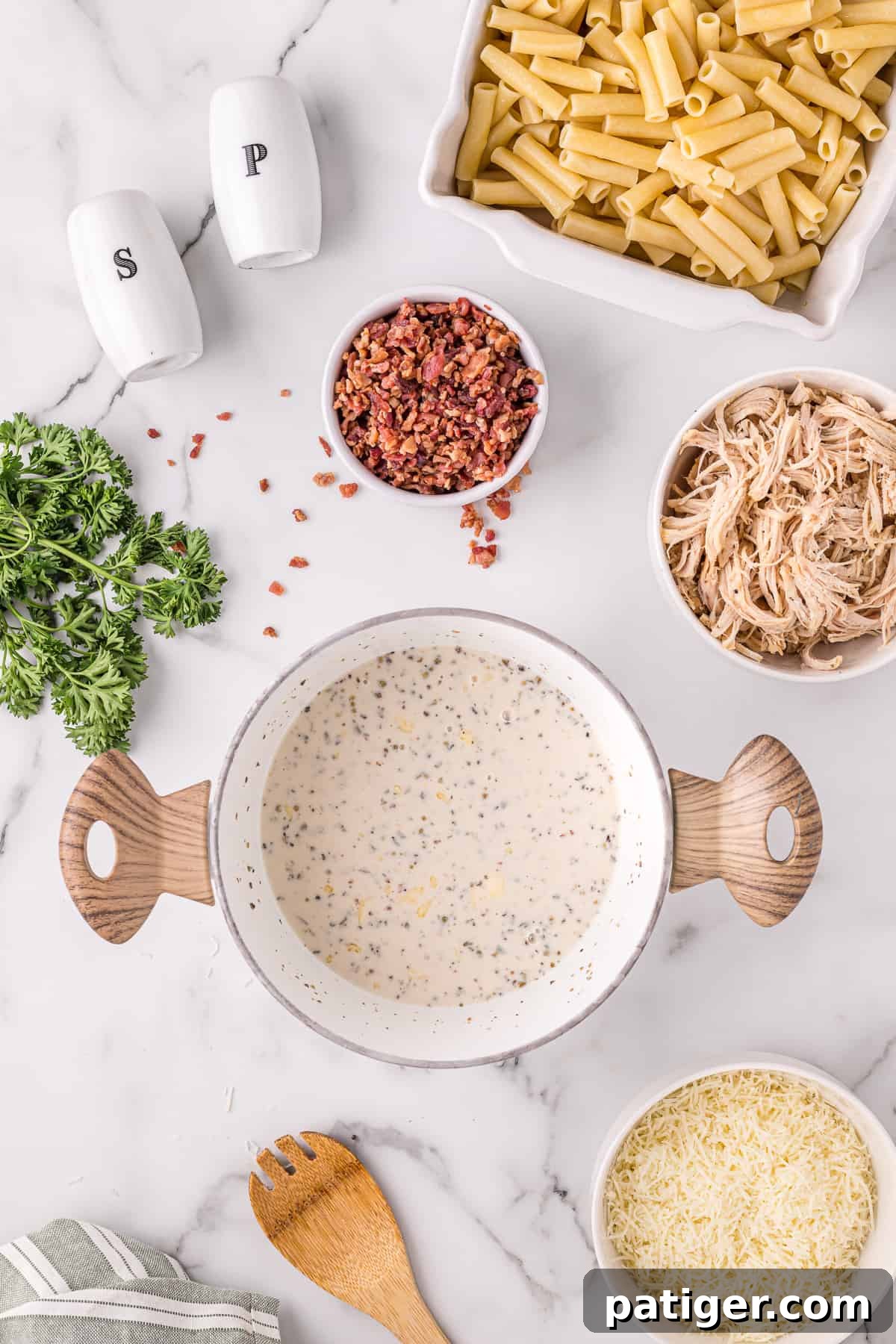 Golden Alfredo Chicken Bake 6 Alfredo sauce simmering in a large pot, surrounded by the other ingredients for the casserole: ziti, chicken, cheese, parsley, and bacon.