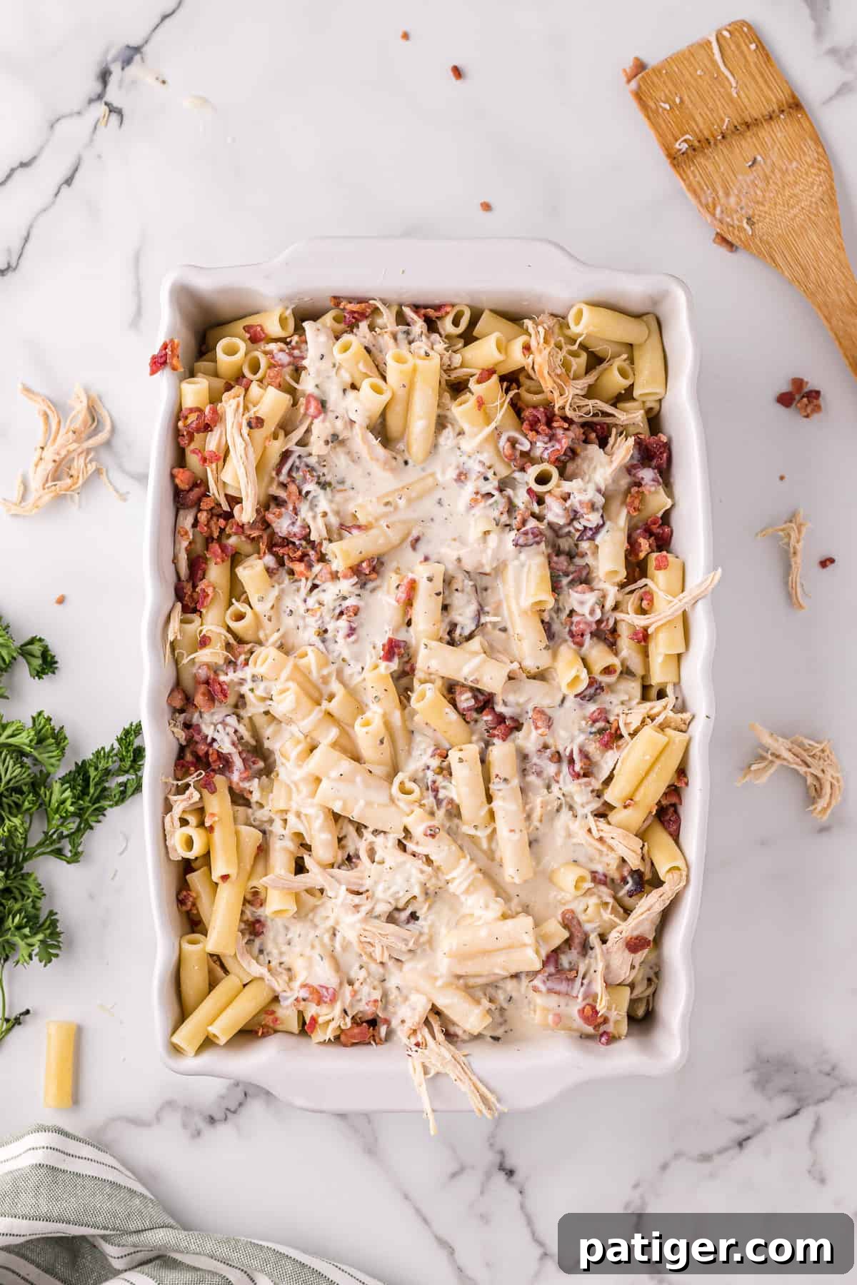 Golden Alfredo Chicken Bake 8 Casserole dish with ziti, crumbled bacon, shredded cooked chicken, and creamy alfredo sauce poured over the top, ready for mixing.