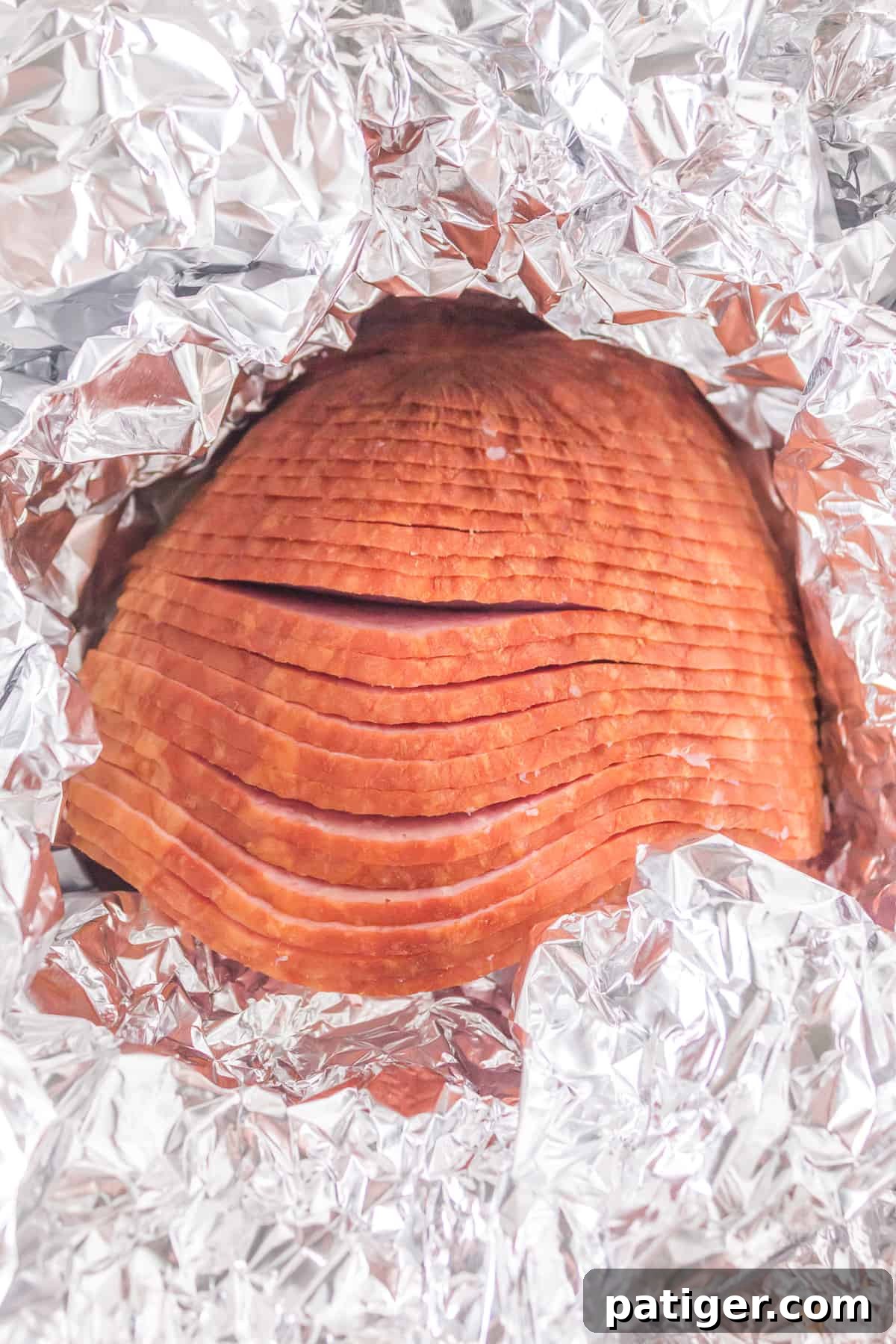 A boneless spiral-cut ham tightly wrapped in aluminum foil, with a portion of the top peeled back to reveal the ham, ready for glazing.