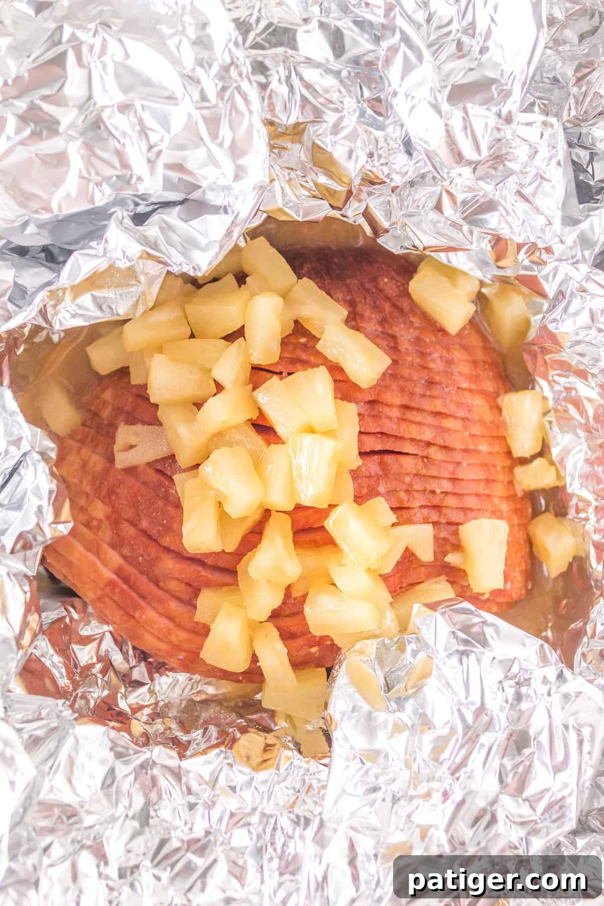 A foil-wrapped ham in the air fryer with the top foil pulled back, revealing the ham generously drizzled with maple pineapple glaze and pineapple chunks.