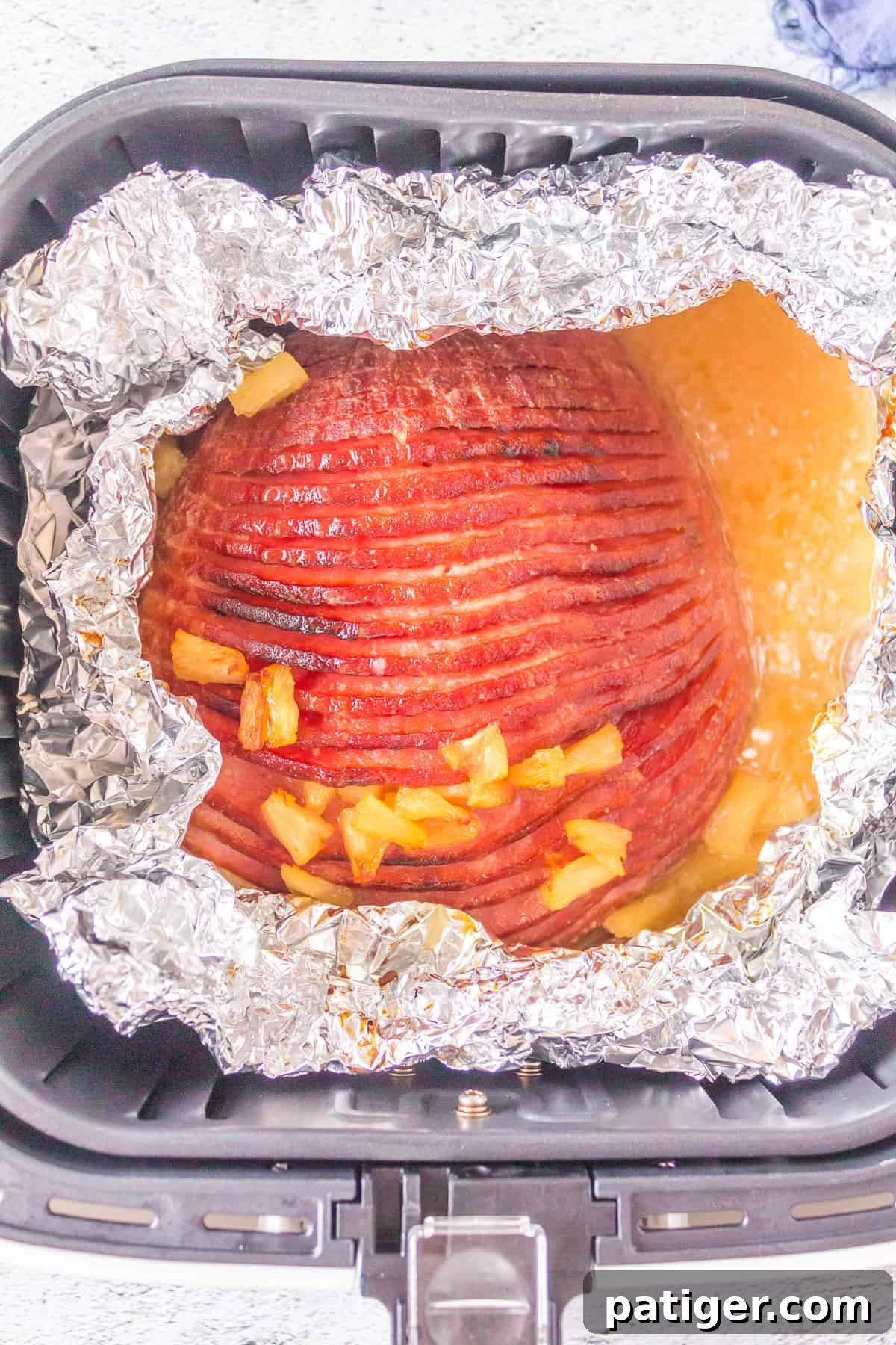A perfectly glazed ham inside an air fryer basket, with the foil pulled back, revealing a browned and caramelized top with cooked pineapple pieces.