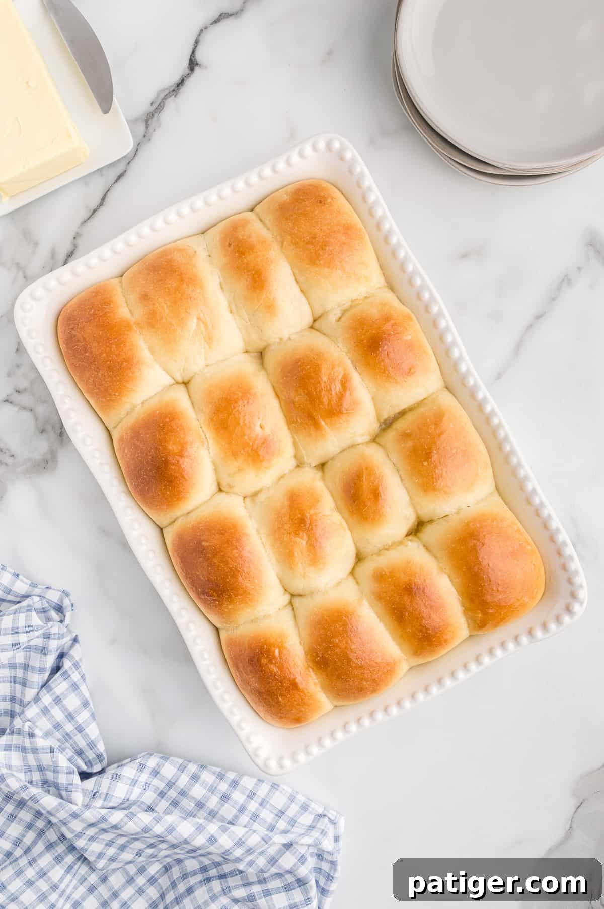 16 golden brown potato dinner rolls in white baking dish with plates, linen, and butter around them