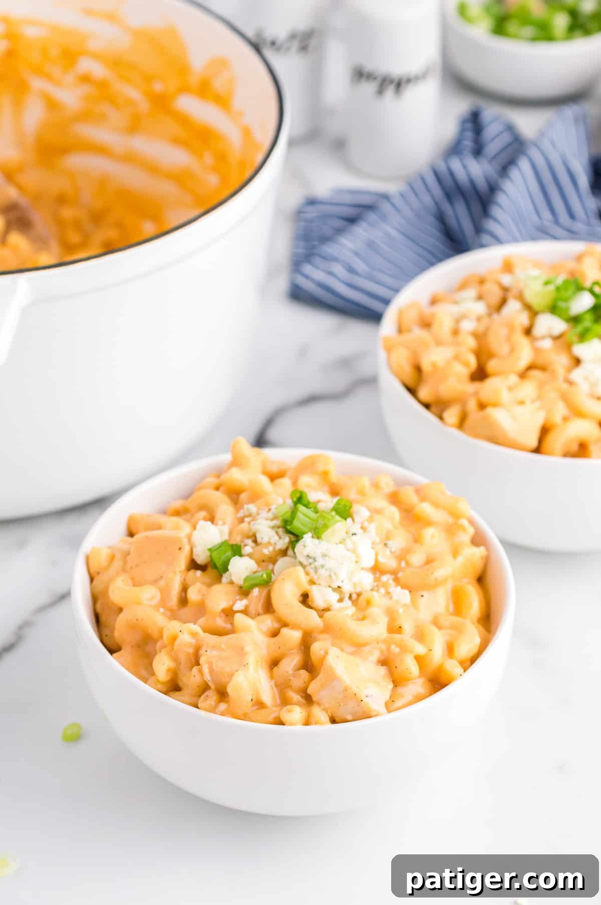 Buffalo Bliss Mac & Cheese 2 A bowl of creamy Buffalo Chicken Mac and Cheese, garnished with blue cheese crumbles and sliced green onions. Another serving and a large pot of pasta are visible in the soft-focus background.