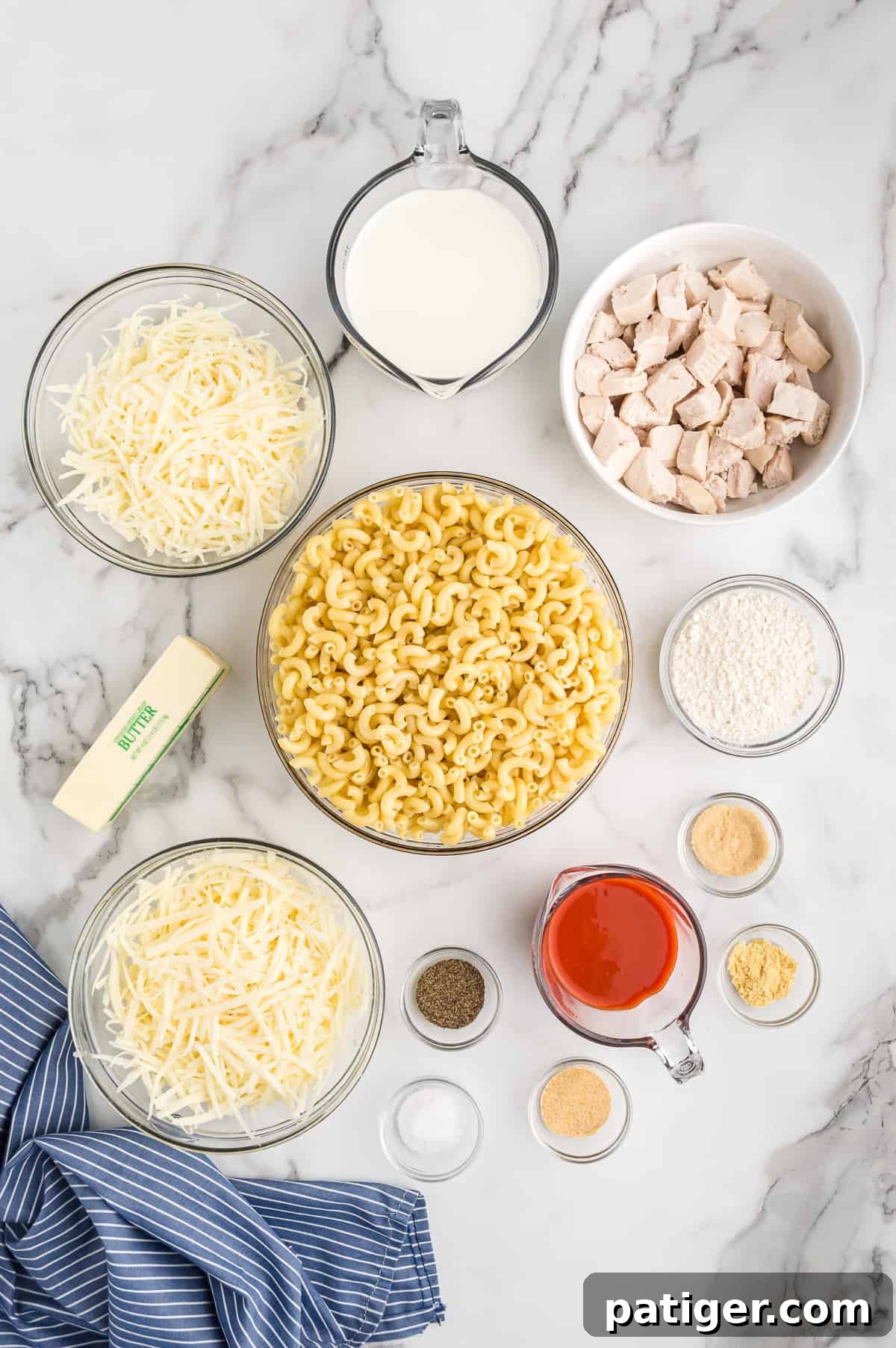 Buffalo Bliss Mac & Cheese 4 A beautiful flat lay of all the fresh ingredients laid out on a marble countertop: a bowl of shredded Monterey Jack, a measuring cup of whole milk, a bowl of cubed cooked chicken, a bowl of all-purpose flour, a measuring cup of buffalo sauce, various small bowls of spices (salt, pepper, onion powder, garlic powder, ground mustard), a second bowl of shredded white cheddar cheese, a stick of unsalted butter, and a bowl of uncooked elbow macaroni.