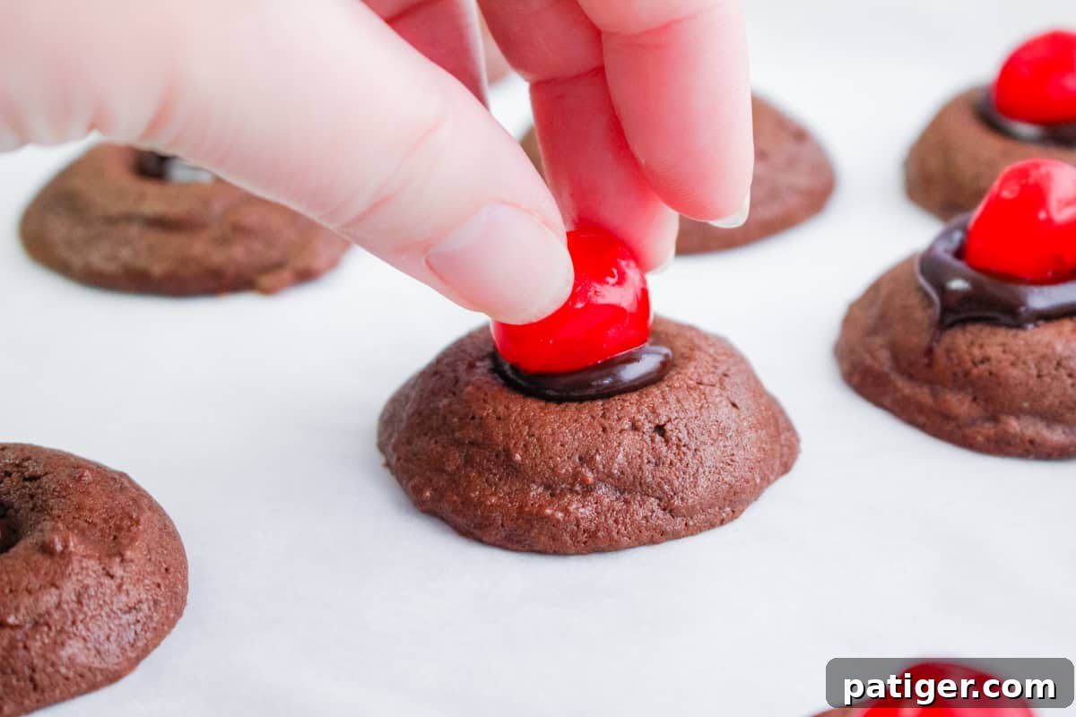 A hand carefully placing a stemless maraschino cherry onto a chocolate thumbprint cookie, which is already filled with glossy ganache.