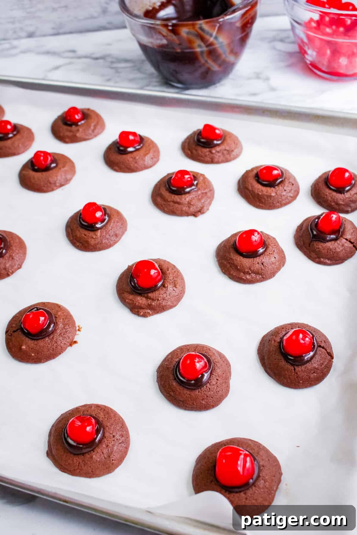 A baking sheet filled with perfectly arranged small chocolate thumbprint cookies, each topped with rich melted chocolate ganache and a bright red maraschino cherry.