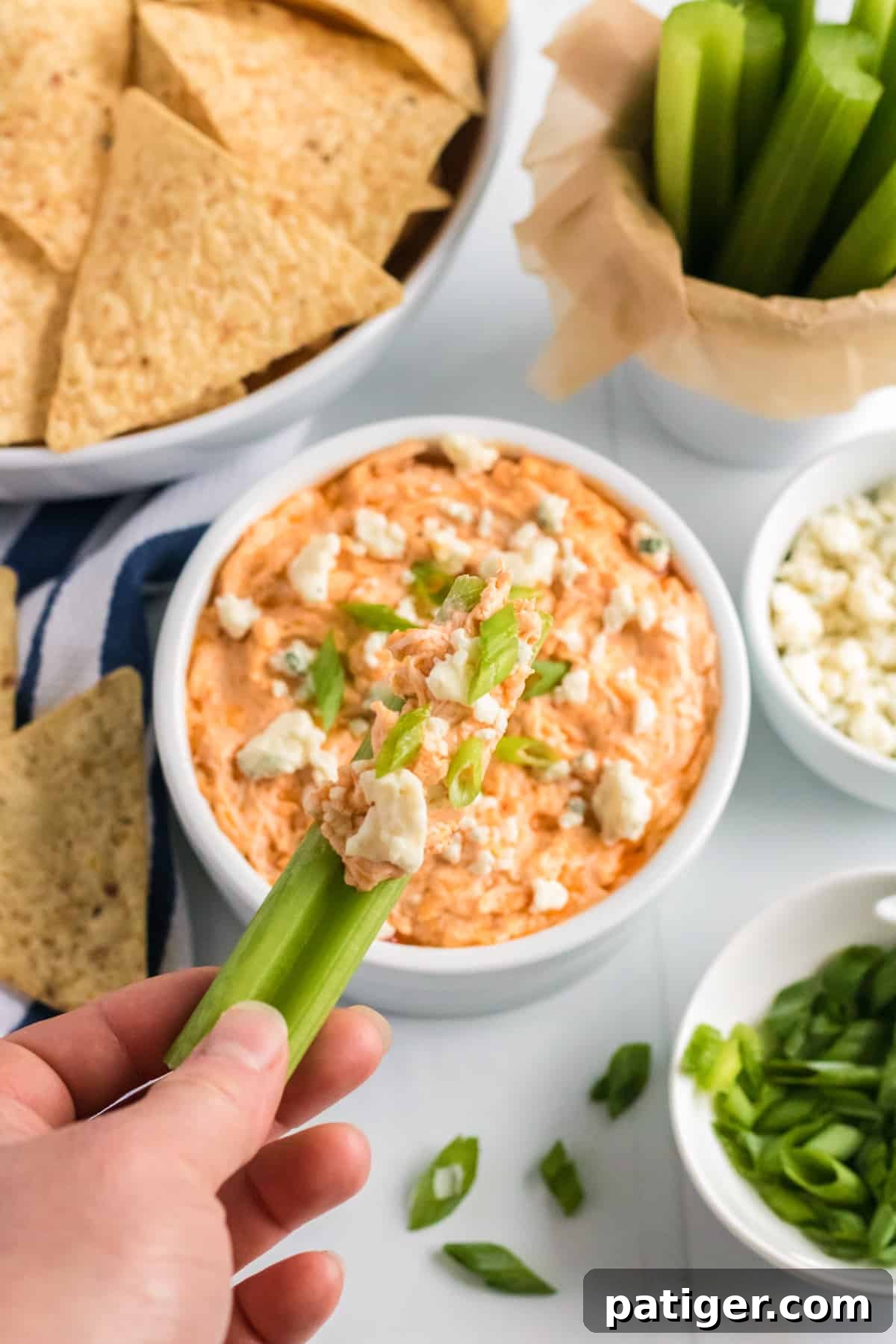 Game Day Crockpot Buffalo Chicken Dip 7 A celery stick being dipped into a bowl of cheesy buffalo chicken dip. In the background, there are tortilla chips, more celery sticks, blue cheese crumbles, and sliced scallions.