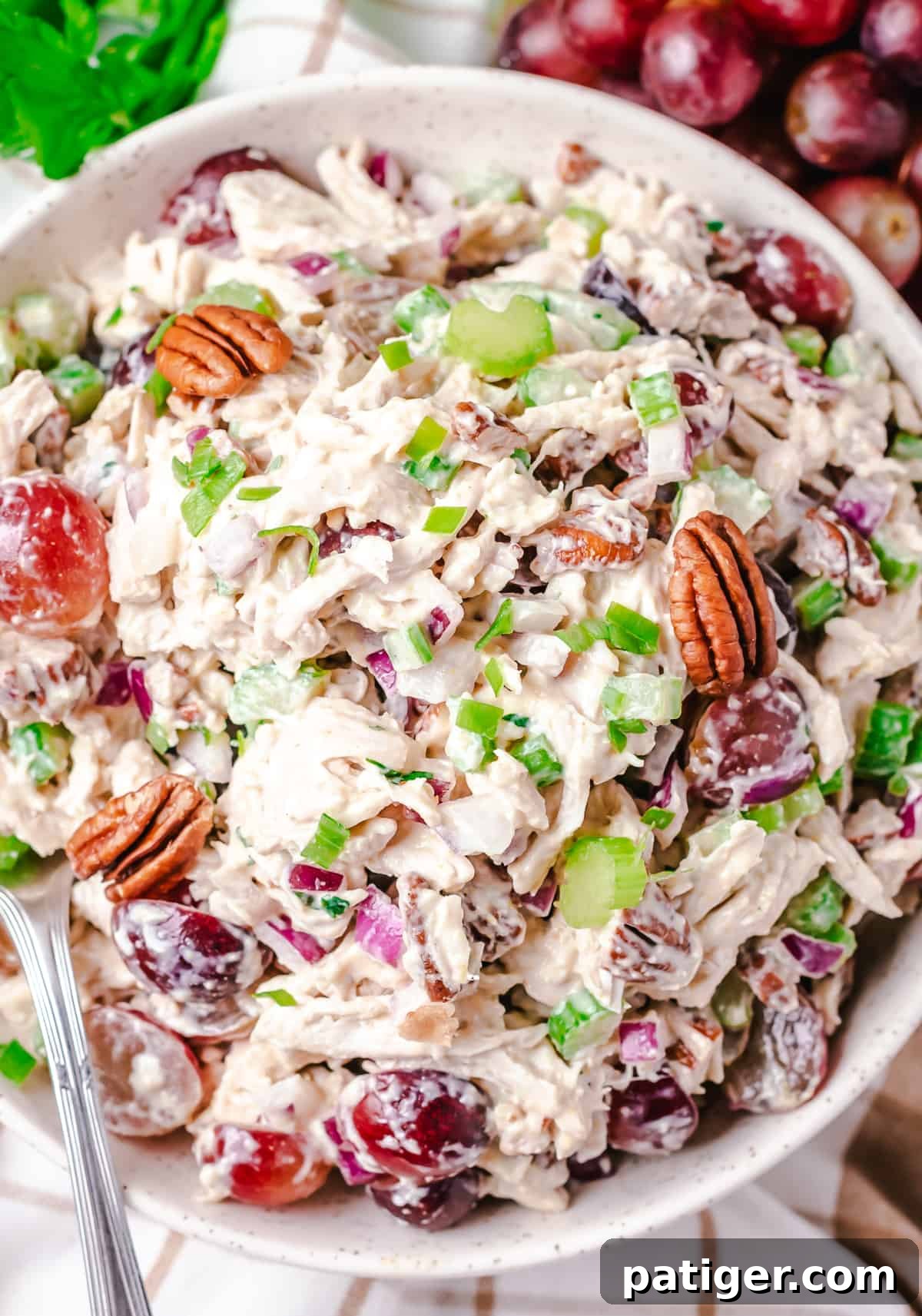 Signature Grape and Pecan Chicken Salad 3 An inviting overhead shot of a large white bowl filled with creamy chicken salad, showcasing its rich texture and colorful mix of ingredients including halved red grapes, chopped pecans, crisp celery, and finely diced red onions, generously garnished with fresh parsley. A scattering of fresh red grapes in the background adds to the visual appeal.