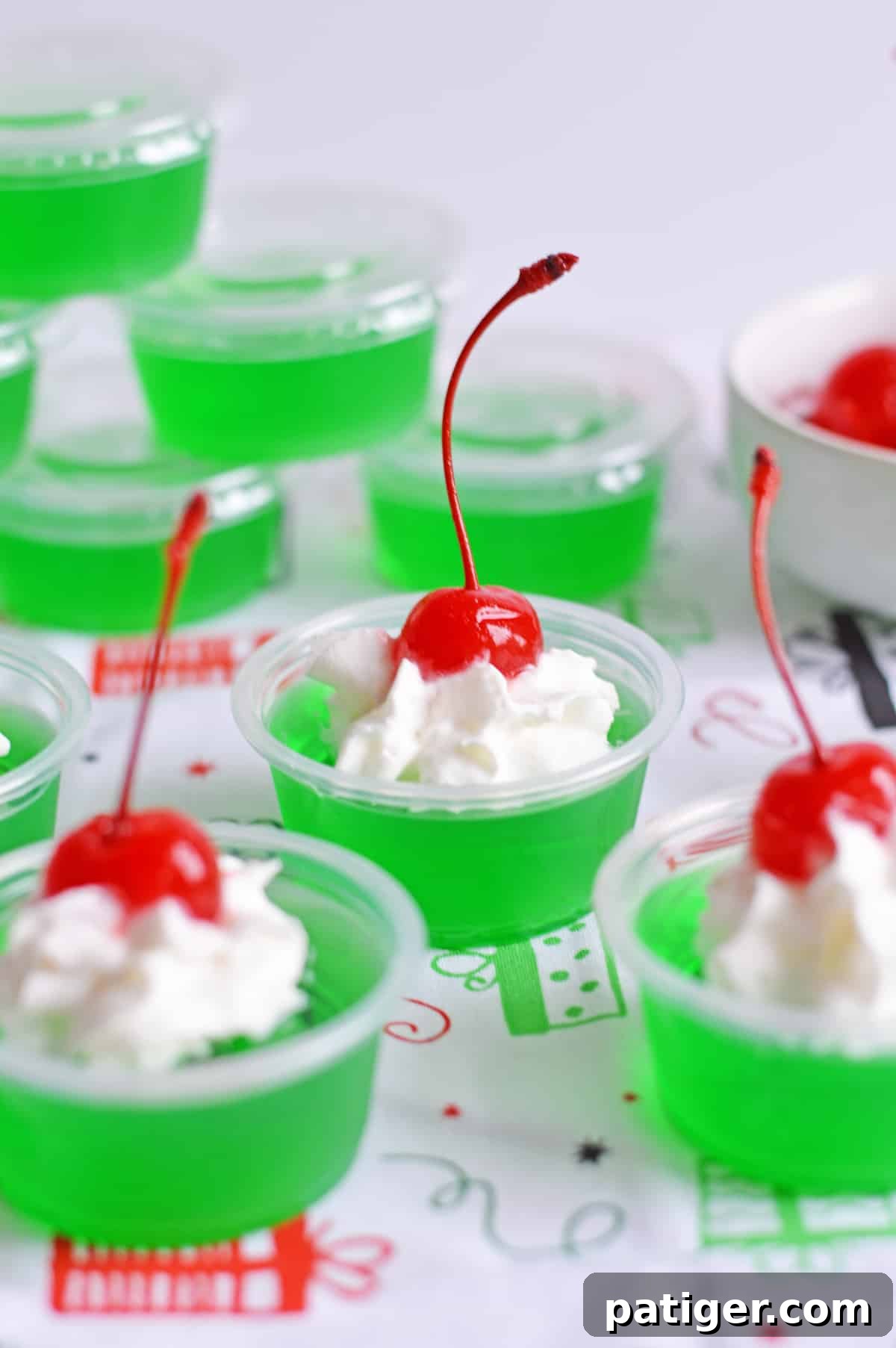 How the Grinch Stole Shots 2 Vibrant green Grinch Jello Shots in small plastic cups, topped with fluffy white whipped cream and a single glistening red maraschino cherry, arranged festively for a Christmas party.