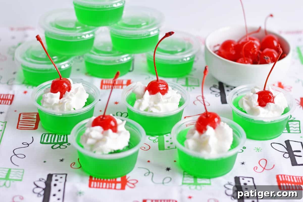 How the Grinch Stole Shots 5 Close-up of perfectly ready-to-serve Christmas Grinch Jello Shots, showcasing the vibrant green jello base generously topped with fluffy white whipped cream and a glistening red maraschino cherry.
