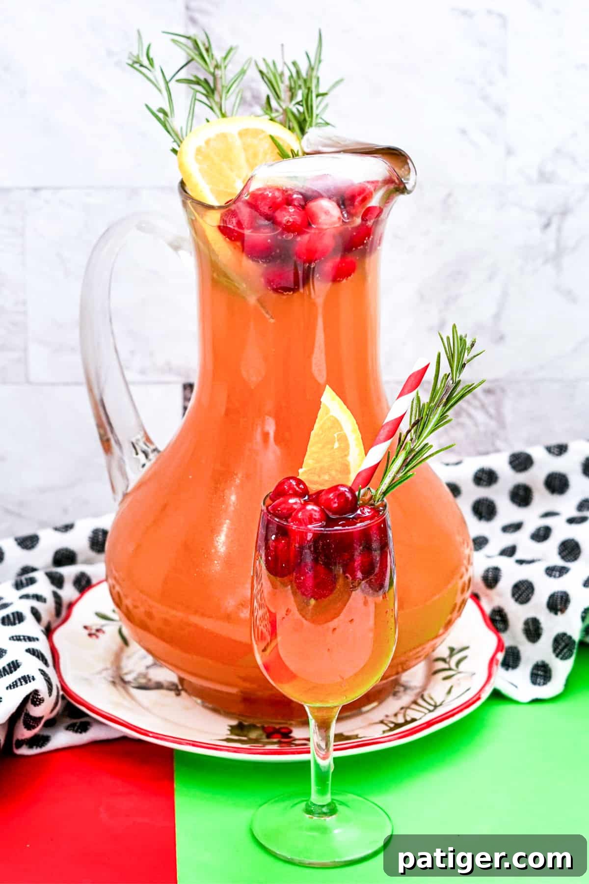 Yuletide Elixir 2 Christmas Punch with vodka and cranberry in a large pitcher garnished with cranberries, orange slice, and rosemary. A glass of the cranberry punch is in front of the pitcher and also garnished with cranberries, orange, and rosemary