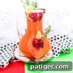 Christmas Punch with vodka and cranberry in a large pitcher garnished with cranberries, orange slice, and rosemary. A glass of the cranberry punch is in front of the pitcher and also garnished with cranberries, orange, and rosemary