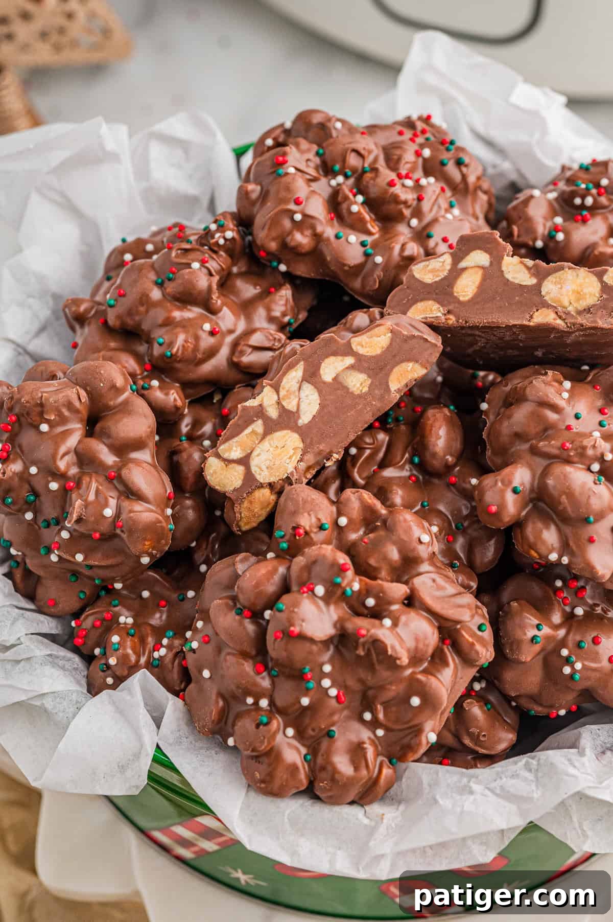 Christmas tin lined with parchment paper and filled with chocolate peanut clusters - "crockpot candy", each topped with sprinkles. One broken cluster reveals whole peanuts inside.