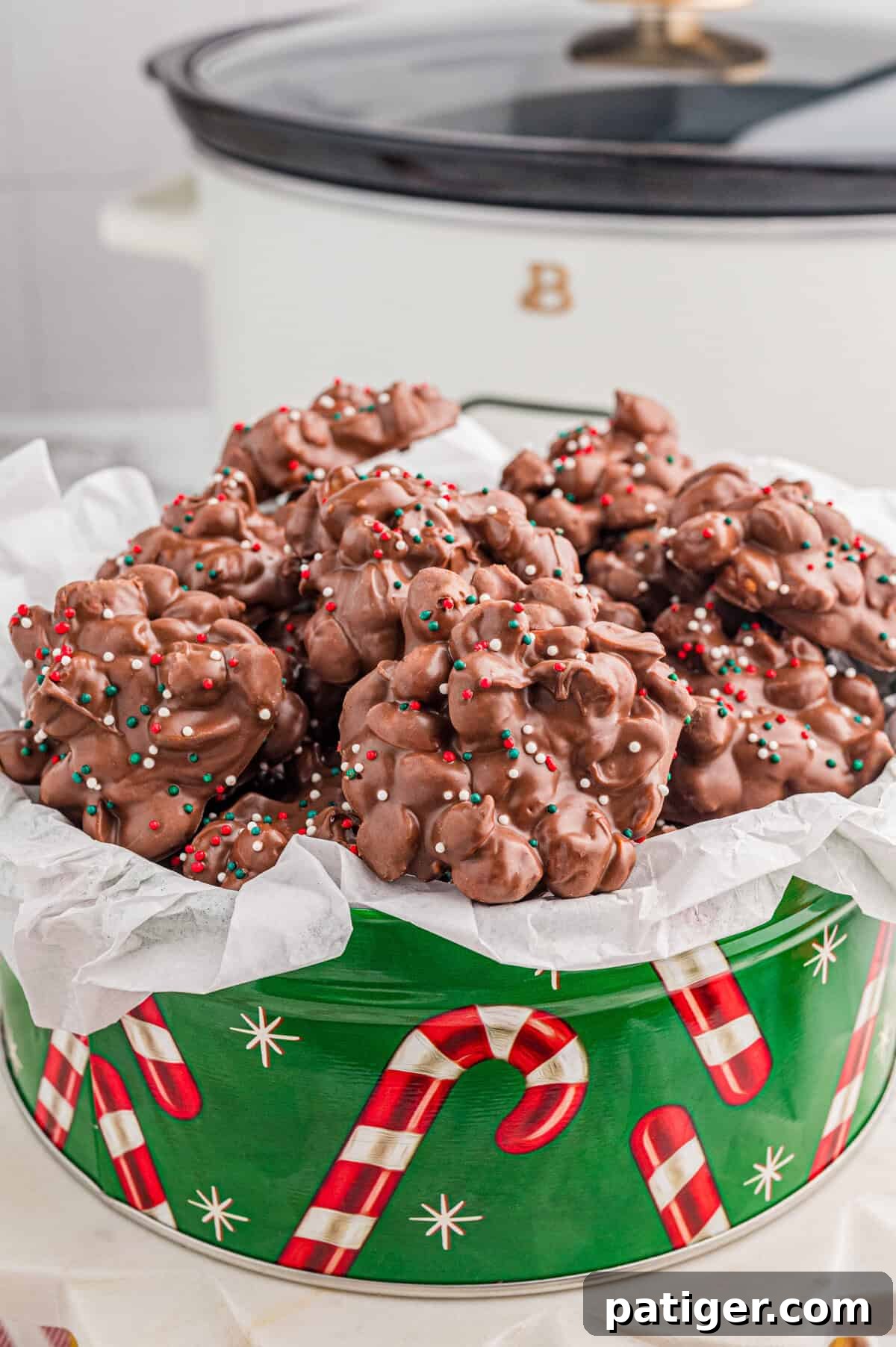 Crock pot Christmas candy in a festive green tin with slow cooker in the background.