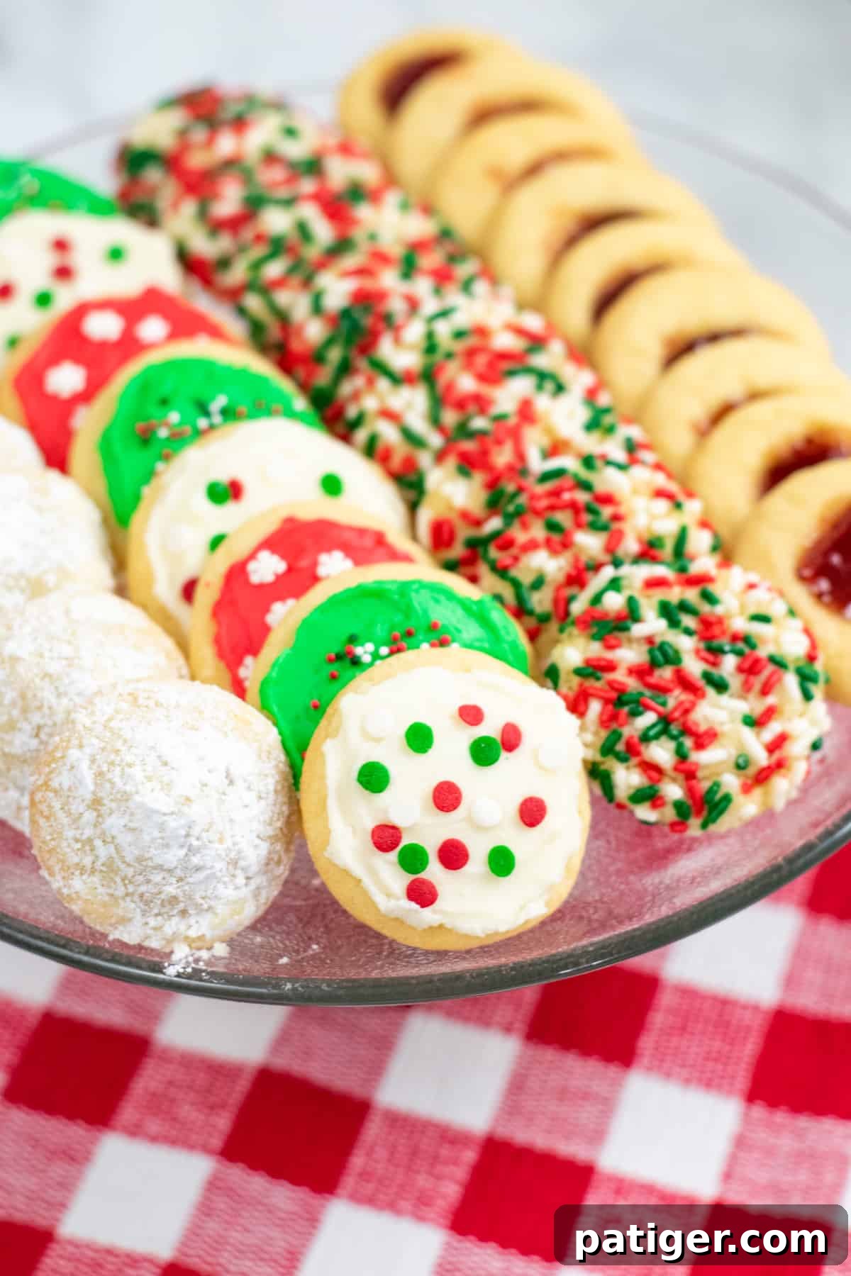 A festive platter showcasing four varieties of Christmas cookies: powdered sugar-dusted hidden kiss cookies, vibrant frosted Christmas cookies, fun sprinkle Christmas cookies, and elegant thumbprint cookies with jam centers.