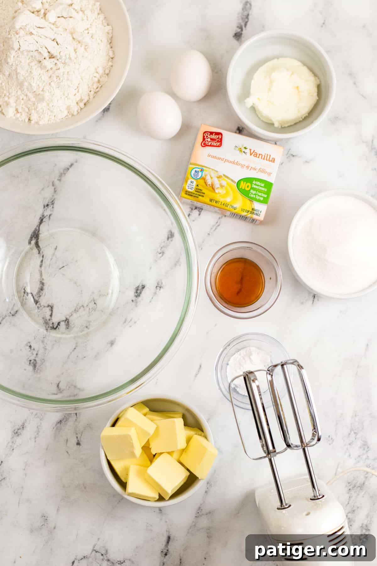 A collection of ingredients laid out for baking vanilla pudding cookies, including a glass mixing bowl, a hand mixer, blocks of butter, bags of flour and sugar, a container of shortening, vanilla pudding mix, eggs, vanilla extract, baking powder, and salt.