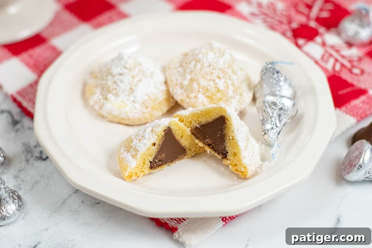 Hidden Kiss Cookies, covered in powdered sugar, on plate with one cut in half to show Herhsey Kiss inside. 