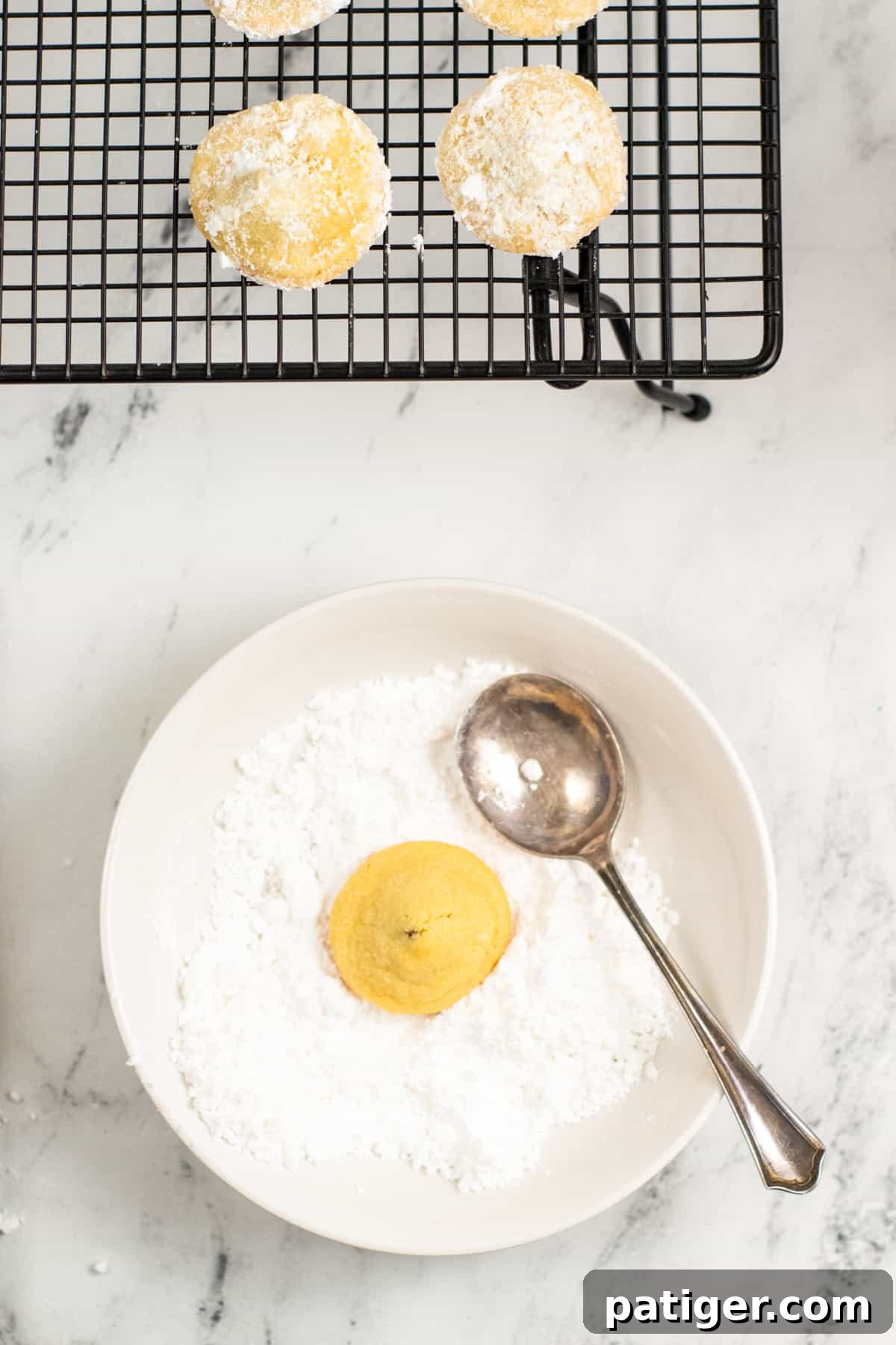Cookie dough in middle of bowl of powdered sugar. A spoon is sticking out of the bowl and wire cooling rack with powdered sugar coated cookies are next to bowl.