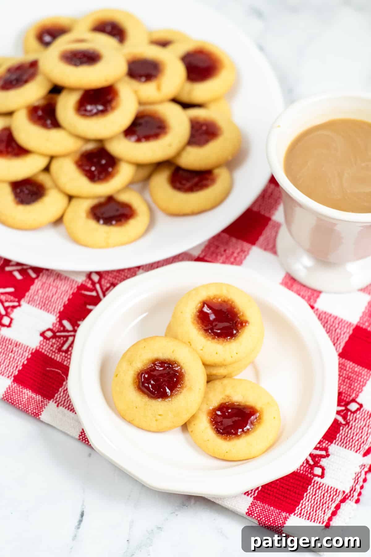 Dreamy Pudding Thumbprints 3 A cozy scene featuring four jam thumbprint cookies alongside a mug of coffee, with a larger platter of cookies in the soft background.