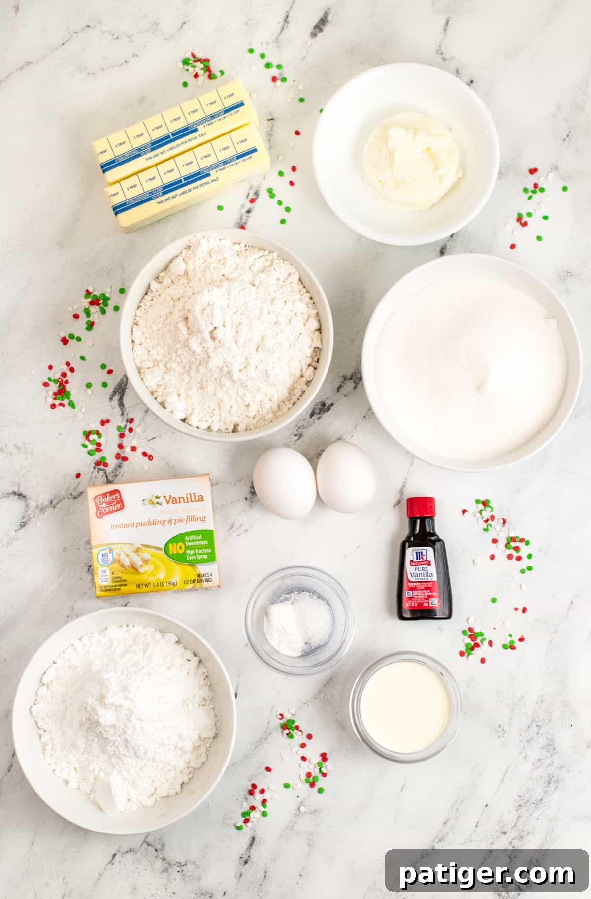 A flat lay of all necessary ingredients for frosted sugar cookies: 2 sticks of butter, shortening, flour, granulated sugar, vanilla pudding mix, vanilla extract, 2 eggs, powdered sugar, baking powder, salt, heavy cream, and Christmas sprinkles on a clean countertop.