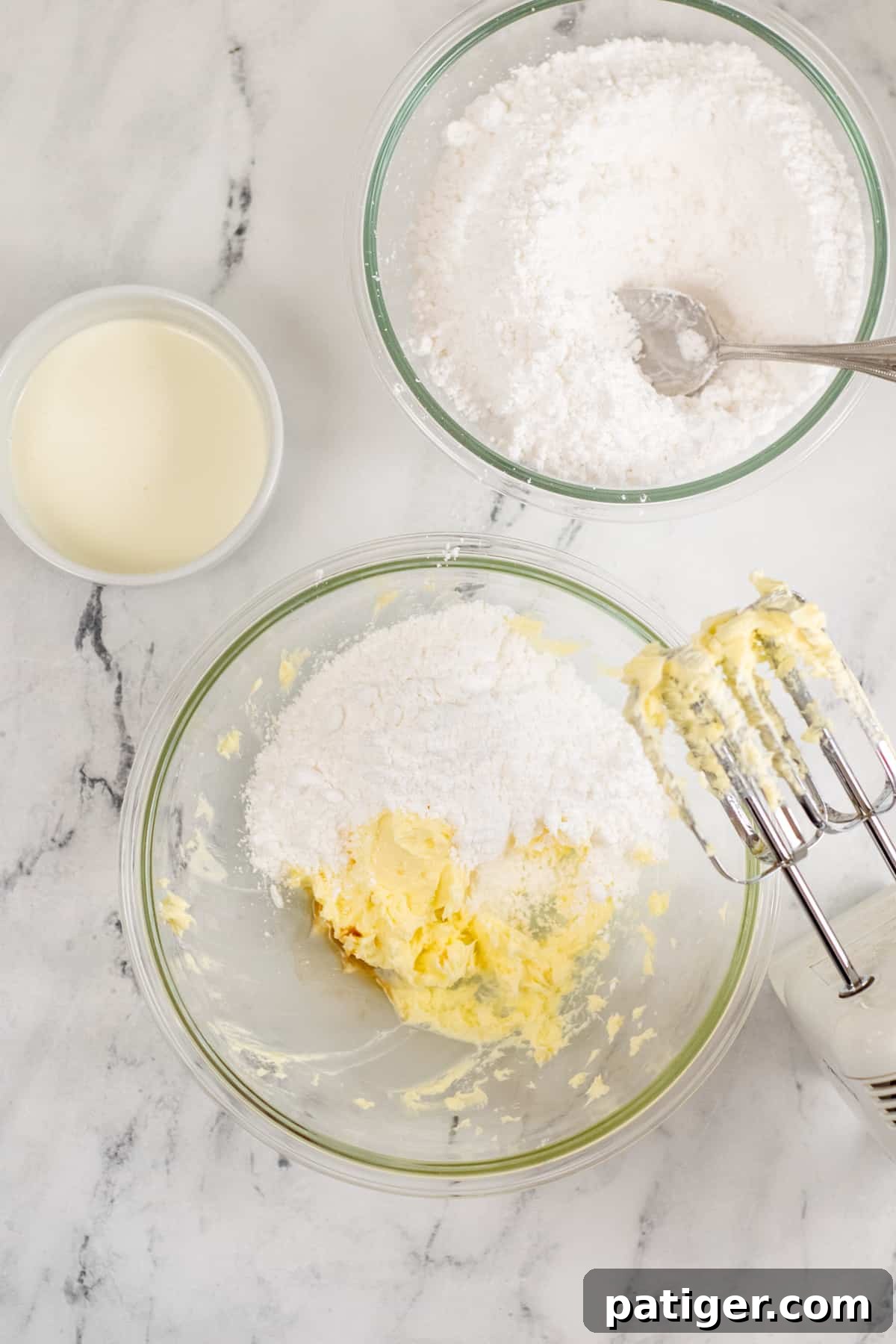 Large glass mixing bowl with creamed butter, powdered sugar, and hand mixer, showing the frosting making process.