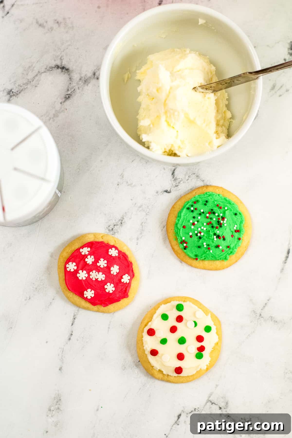 Bowl of frosting, container of sprinkles, and 3 frosted sugar cookies, illustrating the decoration stage.