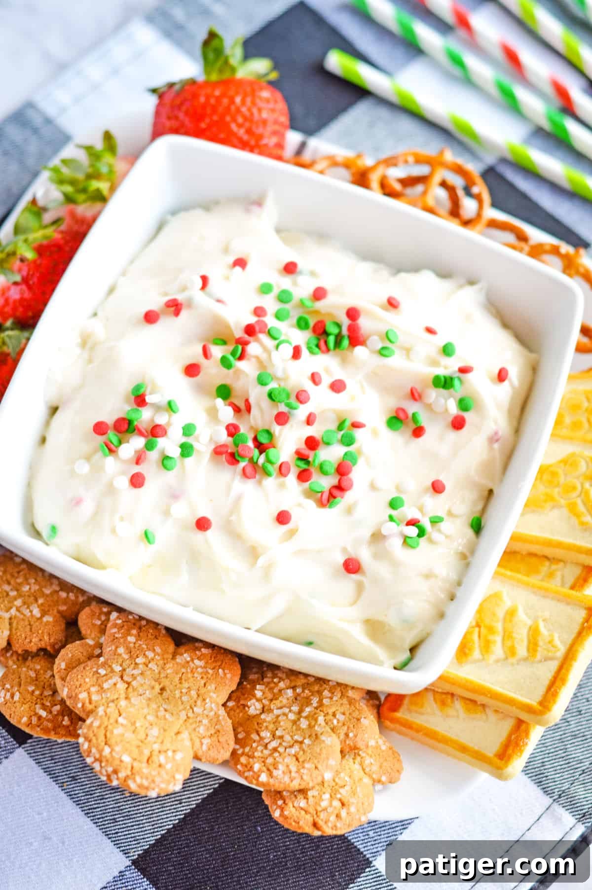 A beautiful white bowl overflowing with Christmas cookie dip, generously topped with colorful Christmas sprinkles. Served alongside are classic Christmas sugar cookies, crunchy pretzels, and fresh strawberries, perfect for dipping.