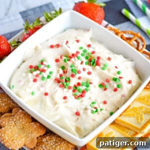A vibrant Christmas dessert dip, served in a festive bowl, with sugar cookies, strawberries, and pretzels artfully arranged around it for dipping.