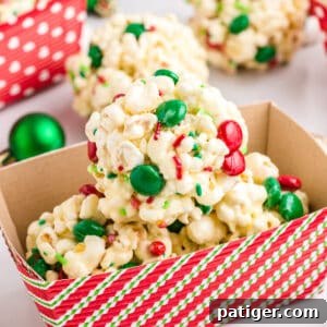 Three round Christmas Popcorn Balls with M&Ms and sprinkles in a paper container for gifting, with more popcorn balls in the background.
