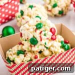 Three round Christmas Popcorn Balls with M&Ms and sprinkles in a paper container for gifting, with more popcorn balls in the background.
