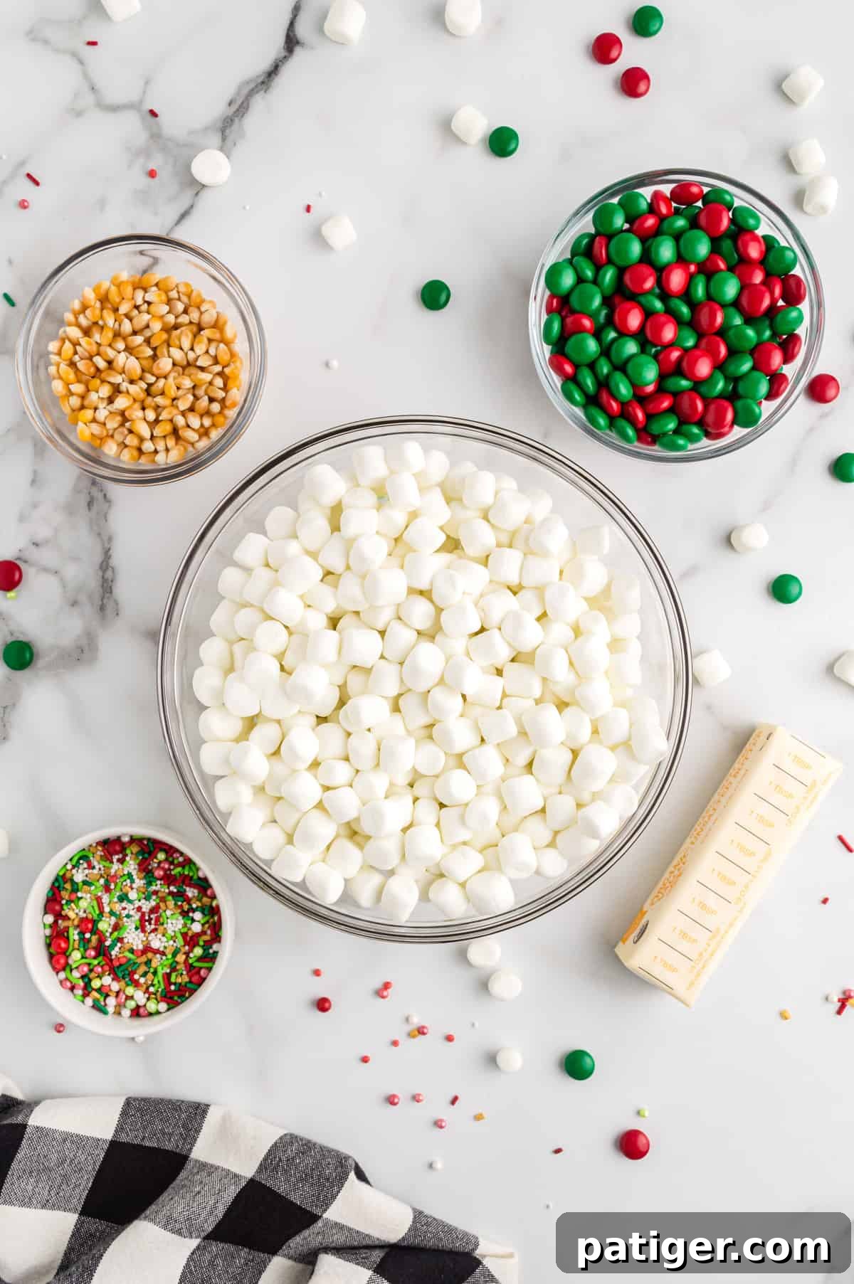 A collection of ingredients for Christmas popcorn balls, including a bowl of unpopped popcorn kernels, mini marshmallows, red and green M&M candies, a stick of butter, and a bowl of festive sprinkles.