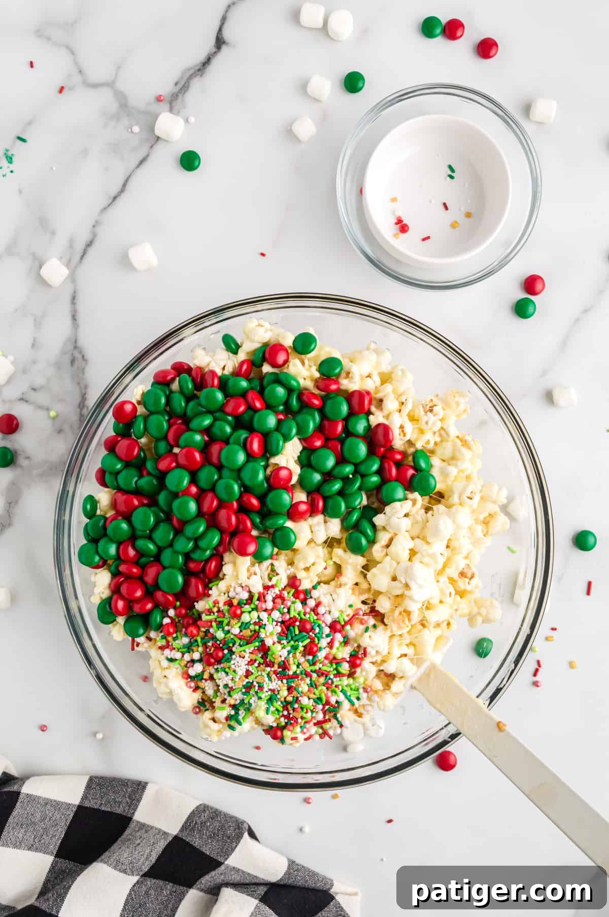 Red and green Christmas M&M candies and festive sprinkles being added to the sticky popcorn mixture, ready to be folded in.
