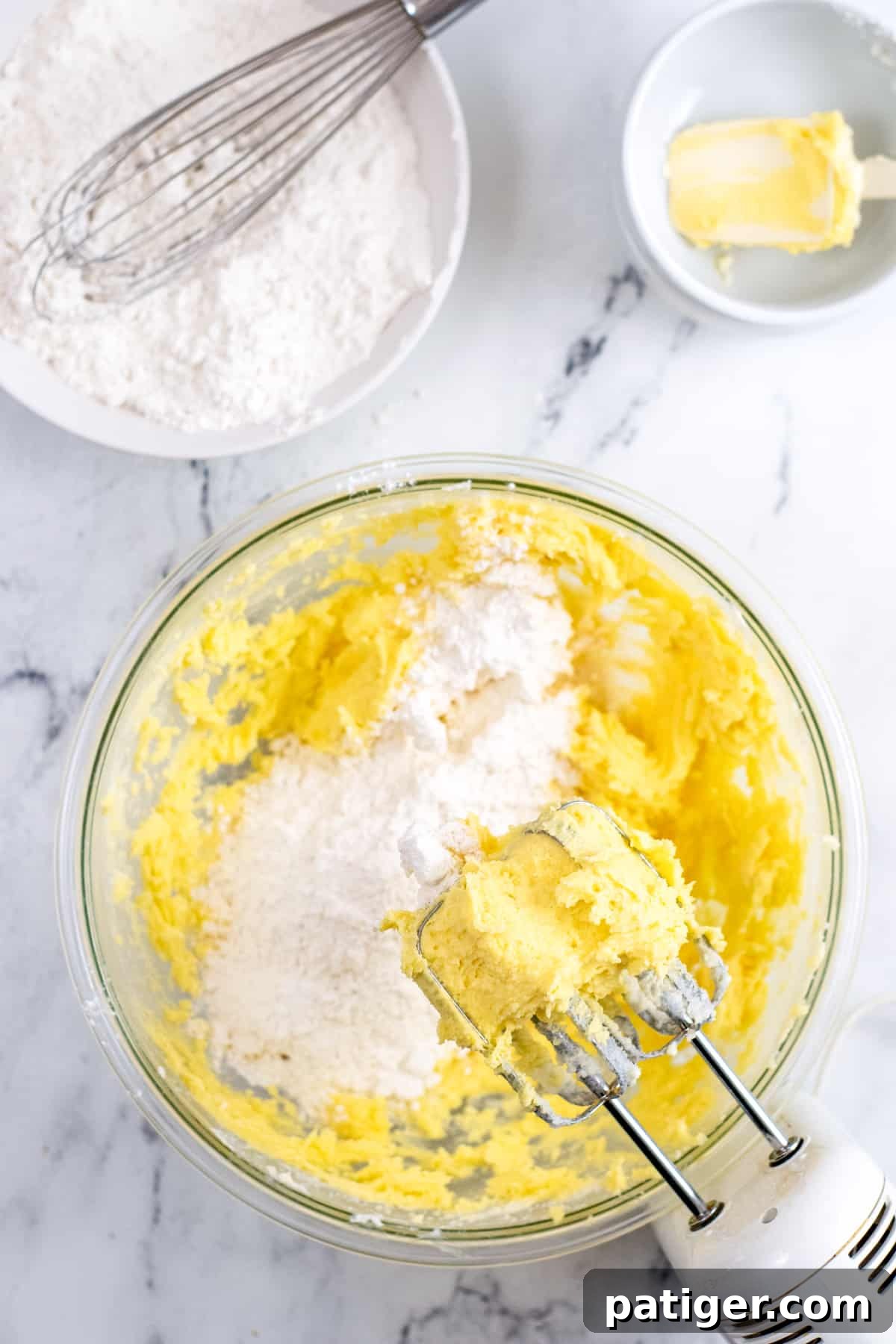 Handheld electric mixer on the side of a large mixing bowl with butter mixture and dry ingredients being gradually added.