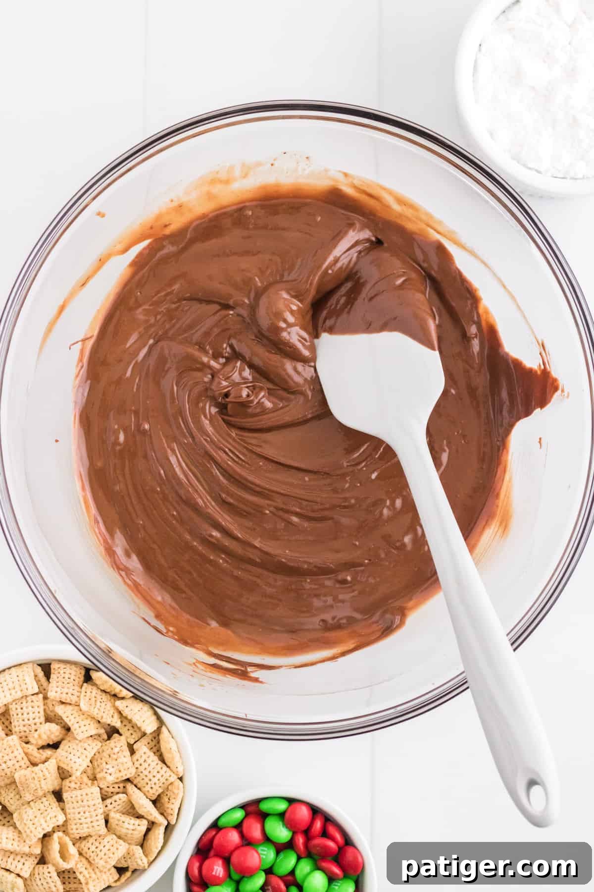 Melted chocolate and peanut butter mixture in large glass bowl. Chex cereal and m&m candies are next to the bowl