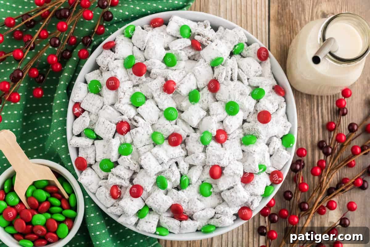Christmas Puppy Chow with M&Ms in a white bowl. Holiday M&Ms, glass of milk, and holiday decor are surrounding the bowl.