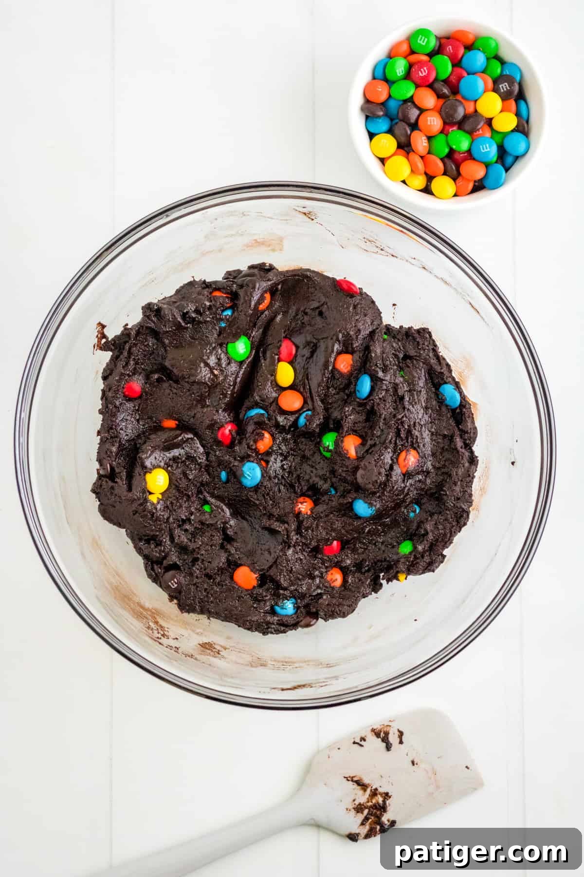 Chocolate M&M Cookie Batter in mixing bowl with spatula and M&Ms next to it.