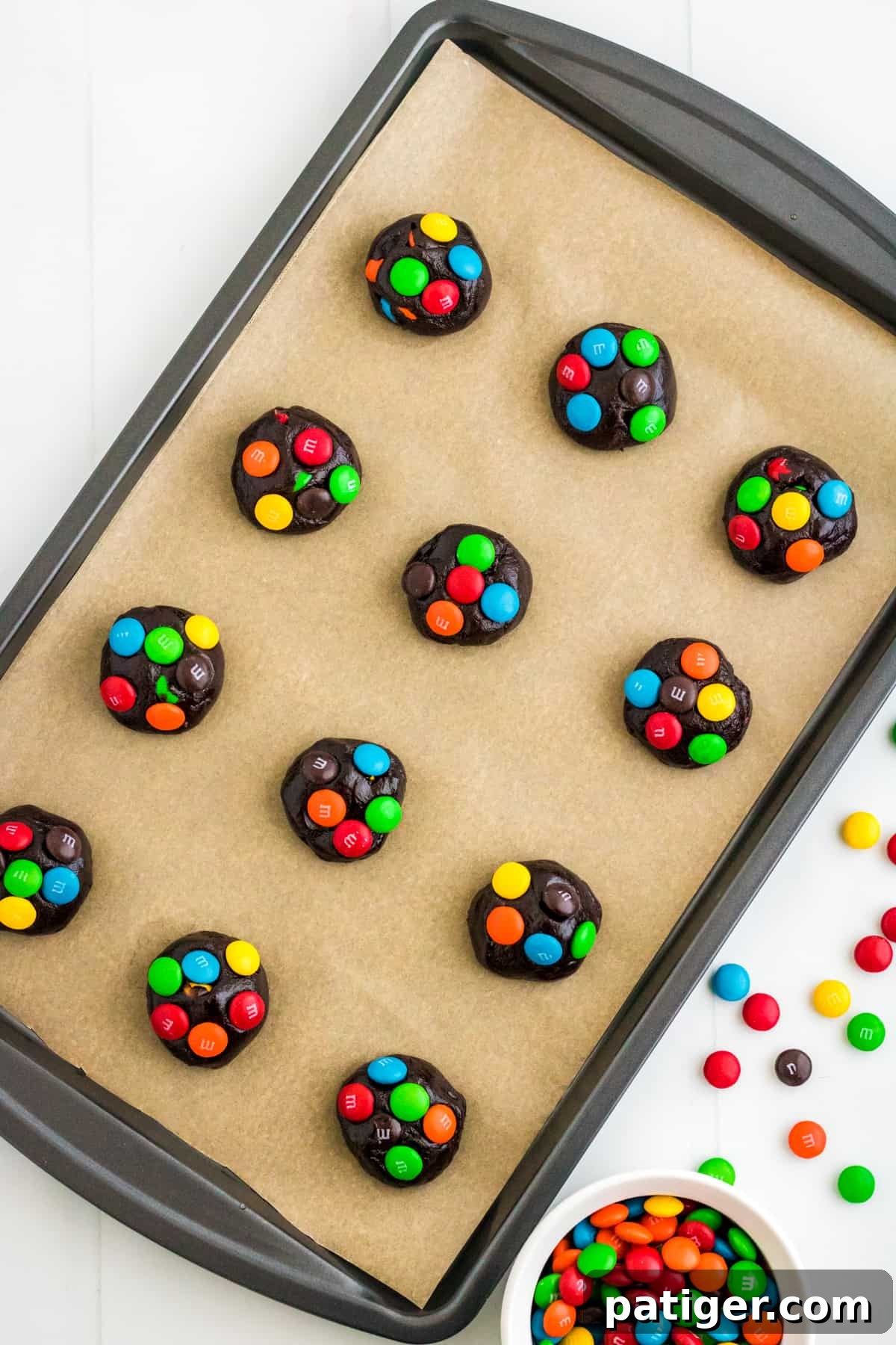 Balls of chocolate cake mix cookie dough with colorful M&Ms pressed into the top on a parchment lined baking sheet.