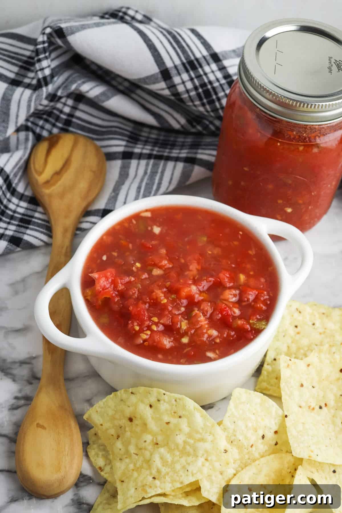 Bowl of homemade Rotel salsa with tortilla chips, cloth napkin, wooden spoons, and jar of homemade salsa in background, showcasing vibrant fresh ingredients for a flavorful dip.