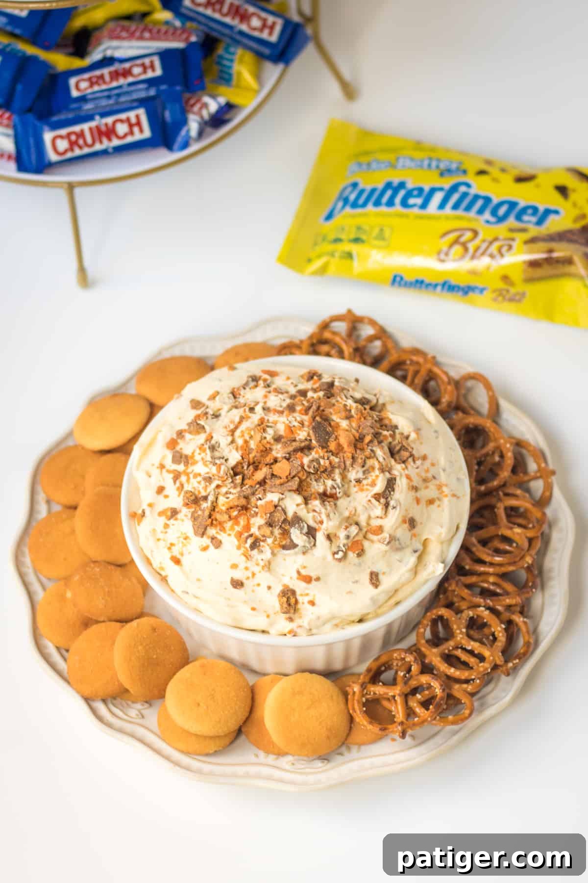 Close-up of a no-bake dessert dip featuring Butterfinger pieces, served with vanilla wafer cookies and pretzel twists for dipping. In the background, a bag of Butterfinger Baking Bits and fun-size Crunch and Butterfinger bars are visible.