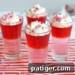 Four Valentine's Day Jello Shots with layers of red and pink gelatin and topped with whipped cream and Valentine's Day Sprinkles