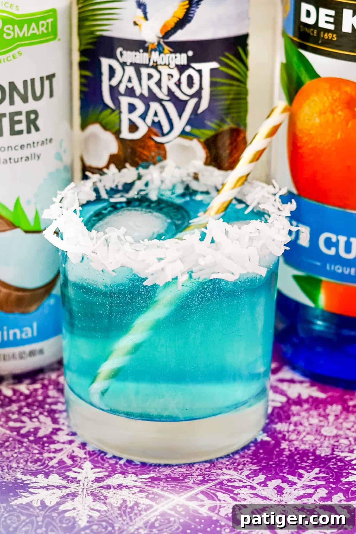 Bright blue Jack Frost cocktail in a rocks glass with a coconut flake rim, with bottles of coconut water, coconut rum, and blue curaçao in the background.