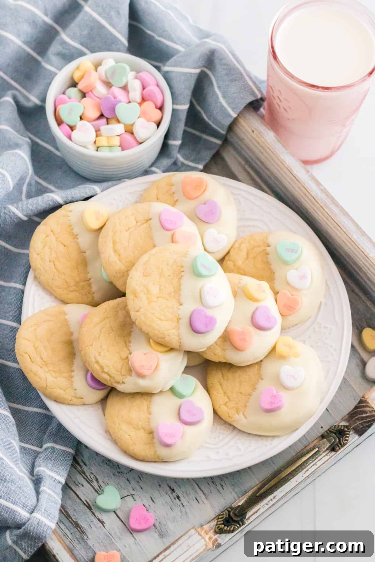 Valentines cake mix cookies dipped in white chocolate and topped with candy hearts. Glass of milk and bowl of conversation heart candies are beside the plate of cookies.